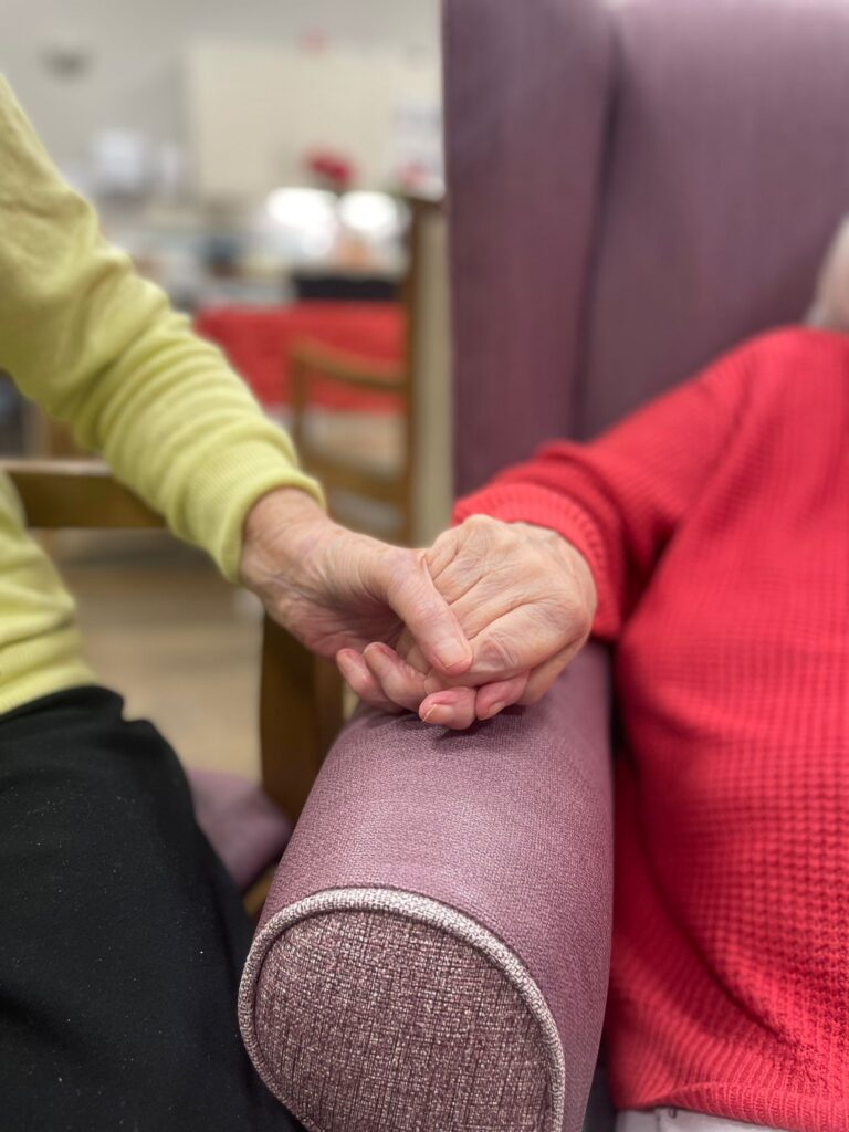 Two people hold hands on the arm of a chair. 
